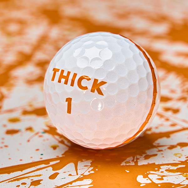 Moon Shot golf ball with an orange THICK 1 logo and the edge of its orange stripe, on white with orange paint splatter